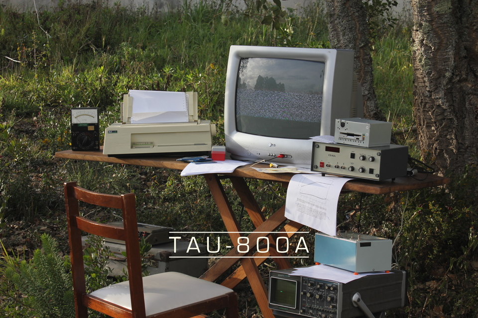 A computer with all manner of retro peripherals, floppy disks, and spare papers, is sitting in a wooden table in the middle of a forest. A caption reads: “TAU-800A”.